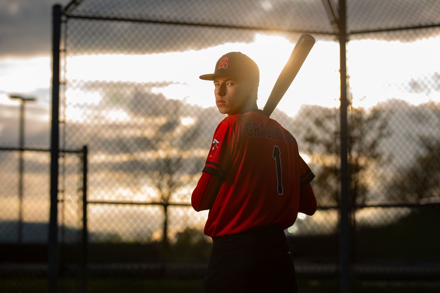 Baseball player in red jersey holding bat at sunset near chain-link fence, dramatic backlit silhouette.