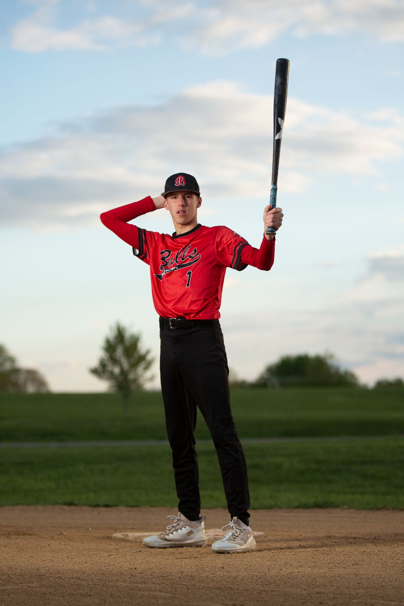 A young baseball player in a red uniform holds a bat raised overhead on a dirt field under a cloudy sky.