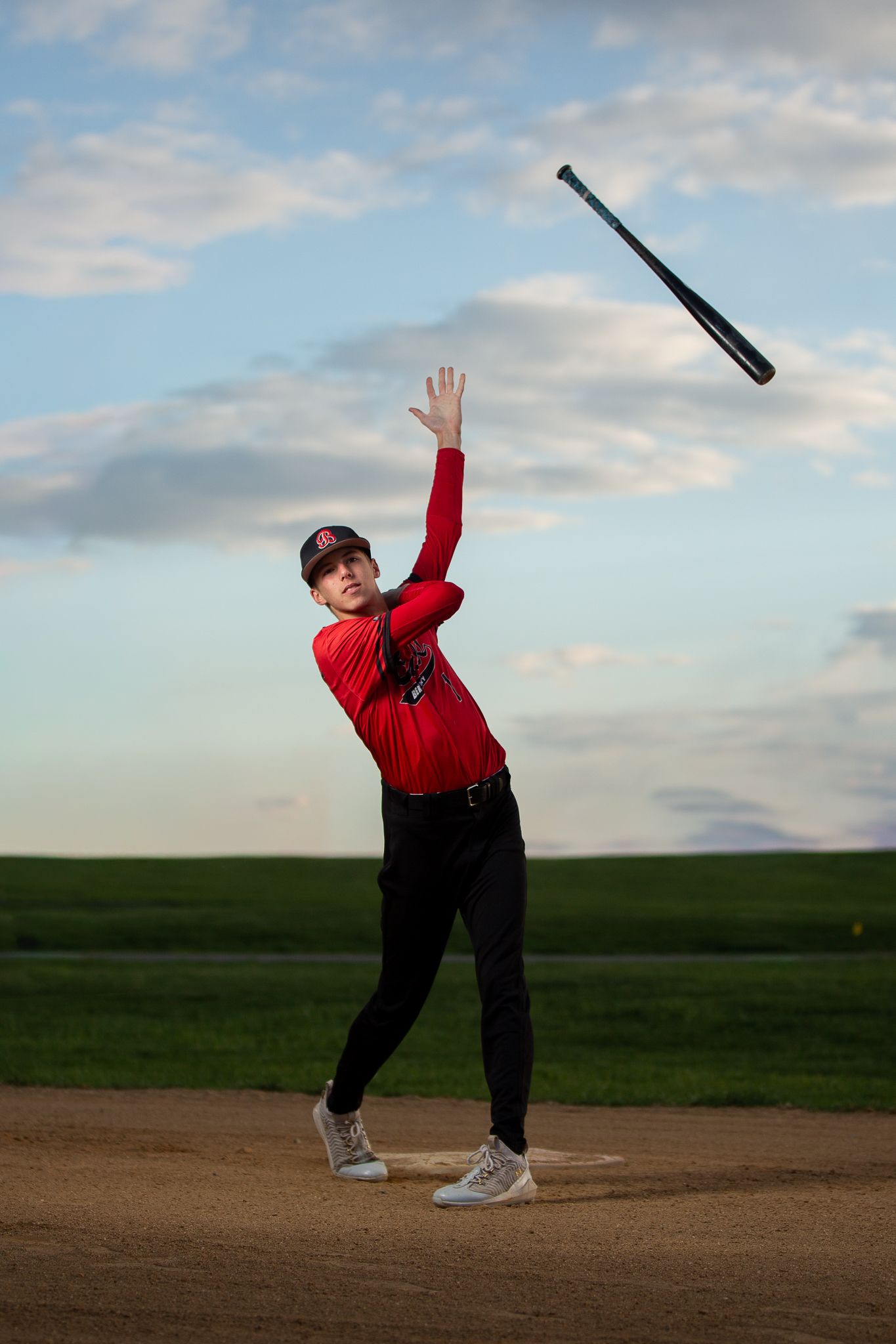 Young baseball player in red uniform tossing a bat into the air on a dirt field with cloudy sky backdrop.