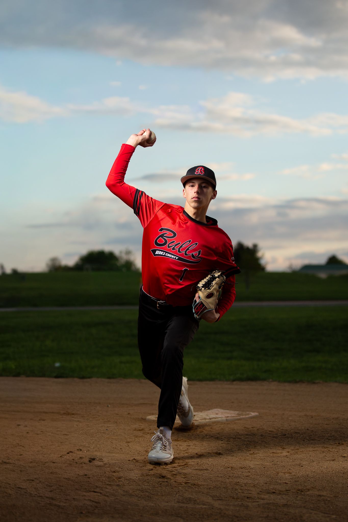 Young baseball pitcher in red Bulls jersey winds up to throw on a dirt mound at dusk.