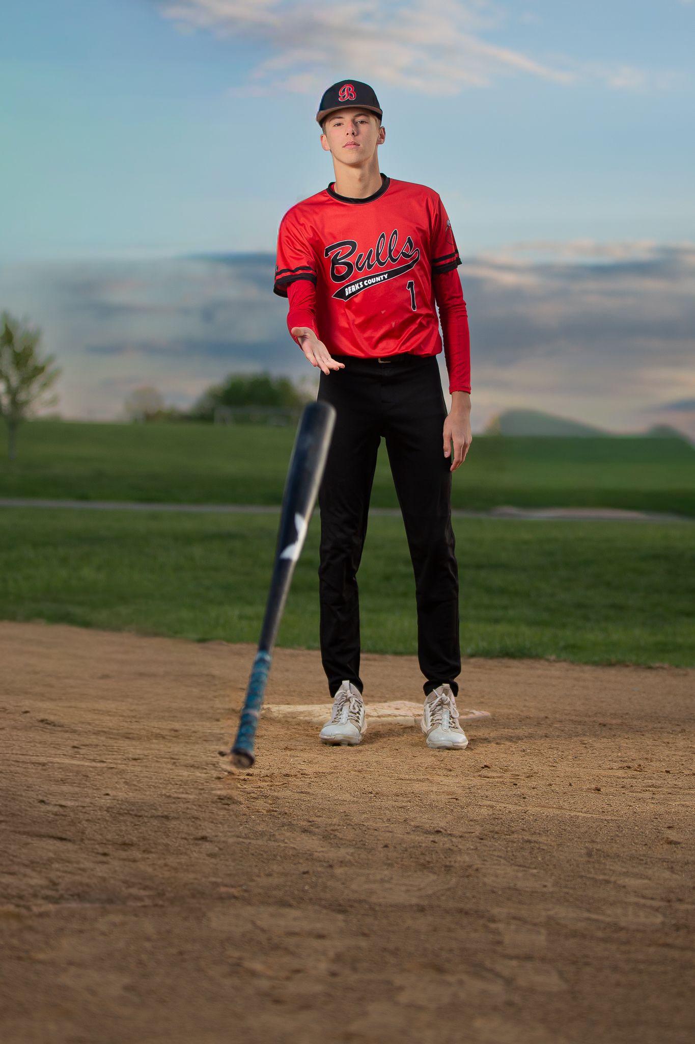 Young baseball player in red Bulls uniform dropping bat on dirt field at dusk.