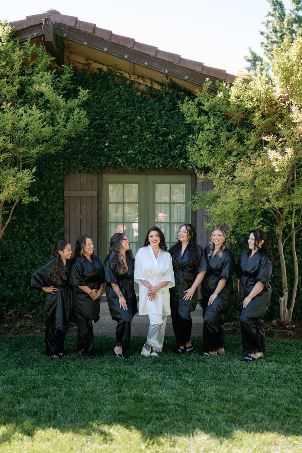 Bride in white robe poses with bridesmaids in black robes on green lawn before ivy-covered building.