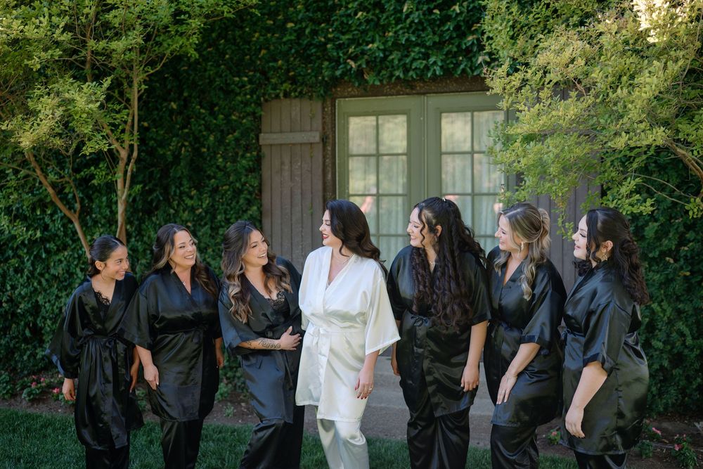 Bride in white robe with bridesmaids in black robes laughing outdoors near ivy-covered doors.