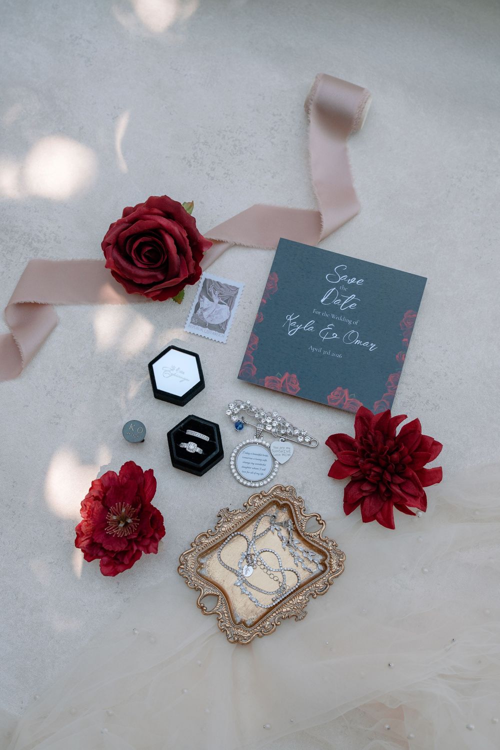 Elegant wedding flat lay with save the date card, engagement ring, red roses, jewelry, and blush ribbon on marble.