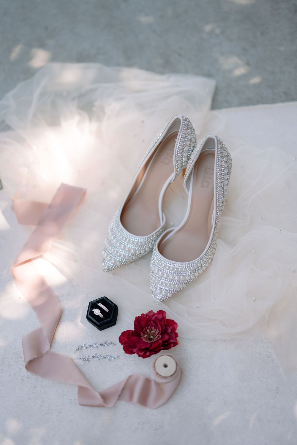 Pearl-embellished bridal flats displayed with pink ribbons, a ring box, and red floral accents on white tulle fabric.