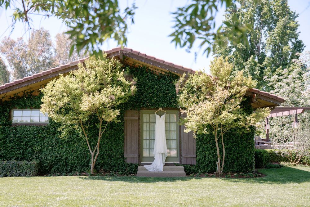 Ivy-covered wedding venue with white bridal gown displayed at entrance, surrounded by lush green trees and manicured lawn.