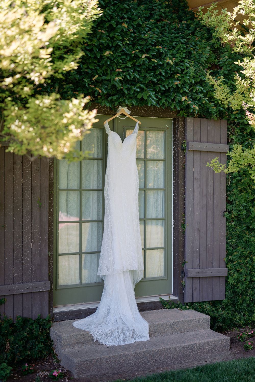 Elegant white lace wedding gown hanging on a wooden hanger outside a charming ivy-covered door.