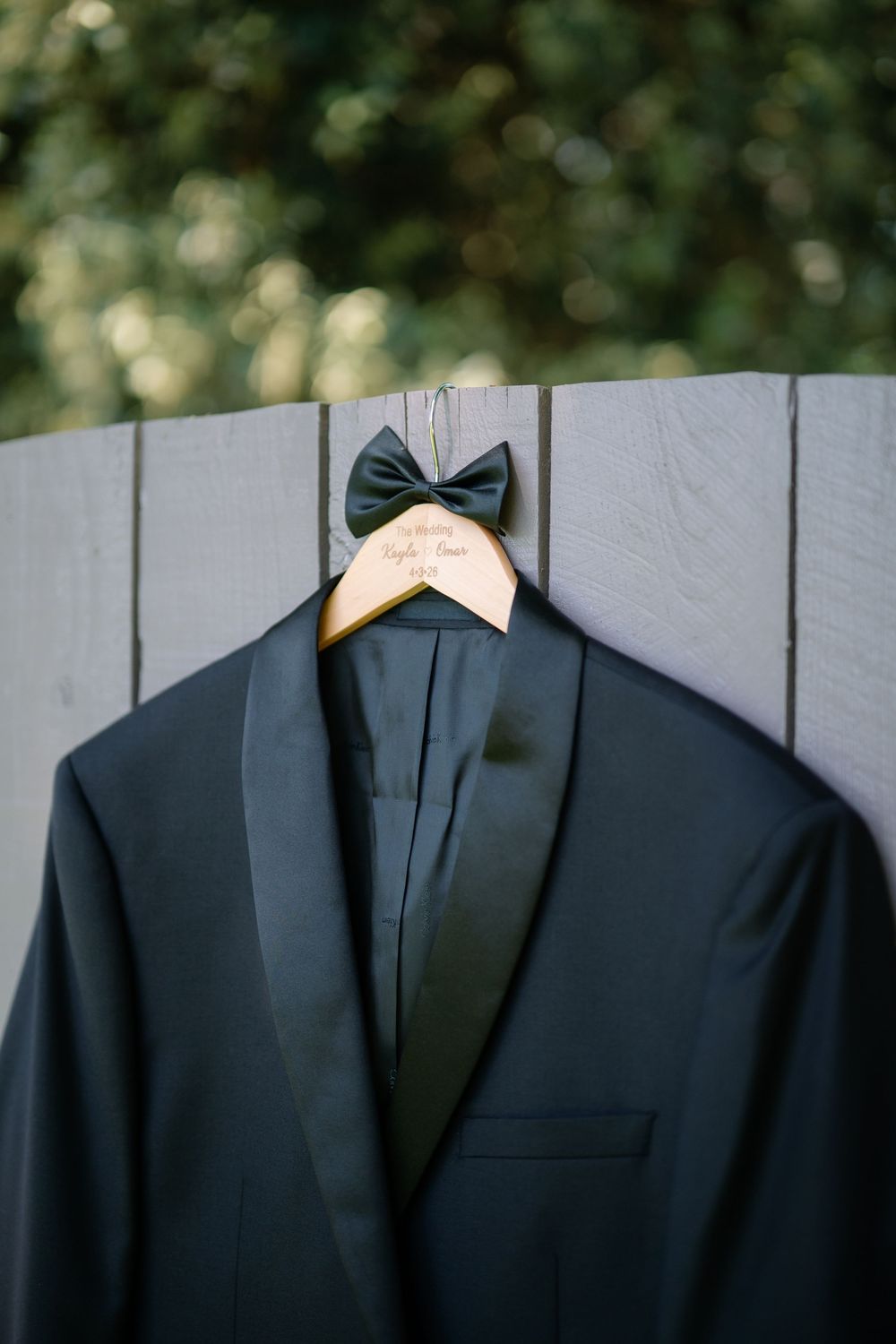 Black tuxedo jacket with dark green bow tie on personalized wooden hanger displayed on rustic fence outdoors.