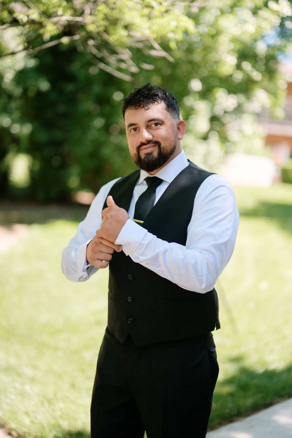 Bearded groom in black vest and white dress shirt adjusting cufflinks outdoors on his wedding day.