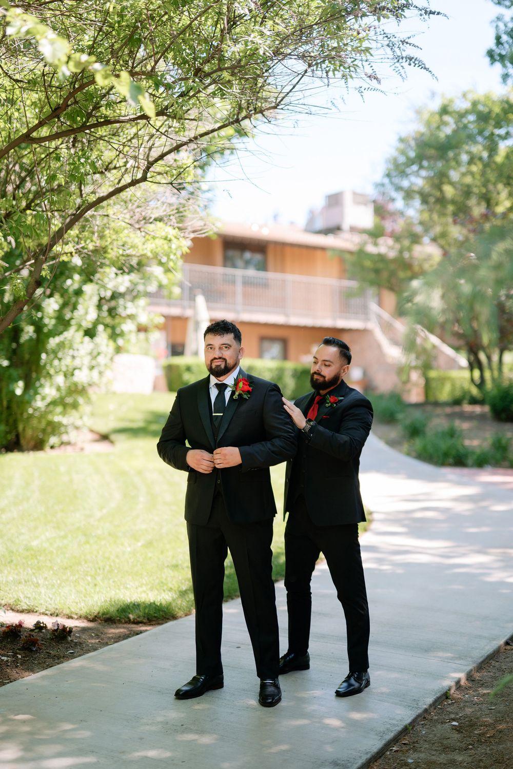Two groomsmen in black suits with red boutonnieres helping each other prepare outdoors.