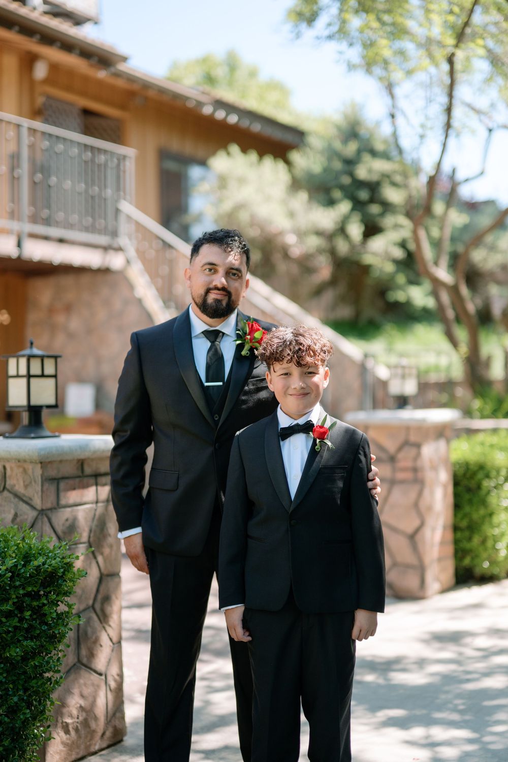 Groom and young ring bearer in matching black suits with red boutonnieres posing outdoors.