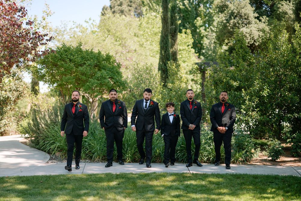 Groomsmen in black tuxedos with red accessories walk together outdoors in a lush garden setting.