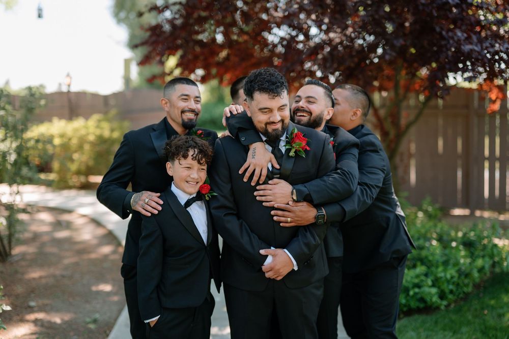 Groom surrounded by smiling groomsmen and a young boy in black suits with red boutonnieres outdoors.