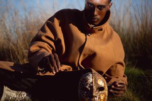 A man in a brown hoodie sits outdoors holding a decorative golden mask among tall grass.