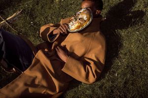 Person in brown coat lying on grass holding a golden mask up to their face in warm sunlight.