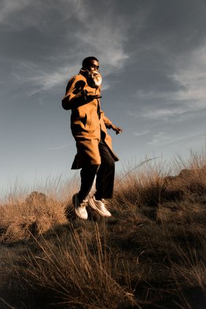 Man in brown coat and white sneakers jumping on a grassy hill at golden hour with dramatic cloudy sky.