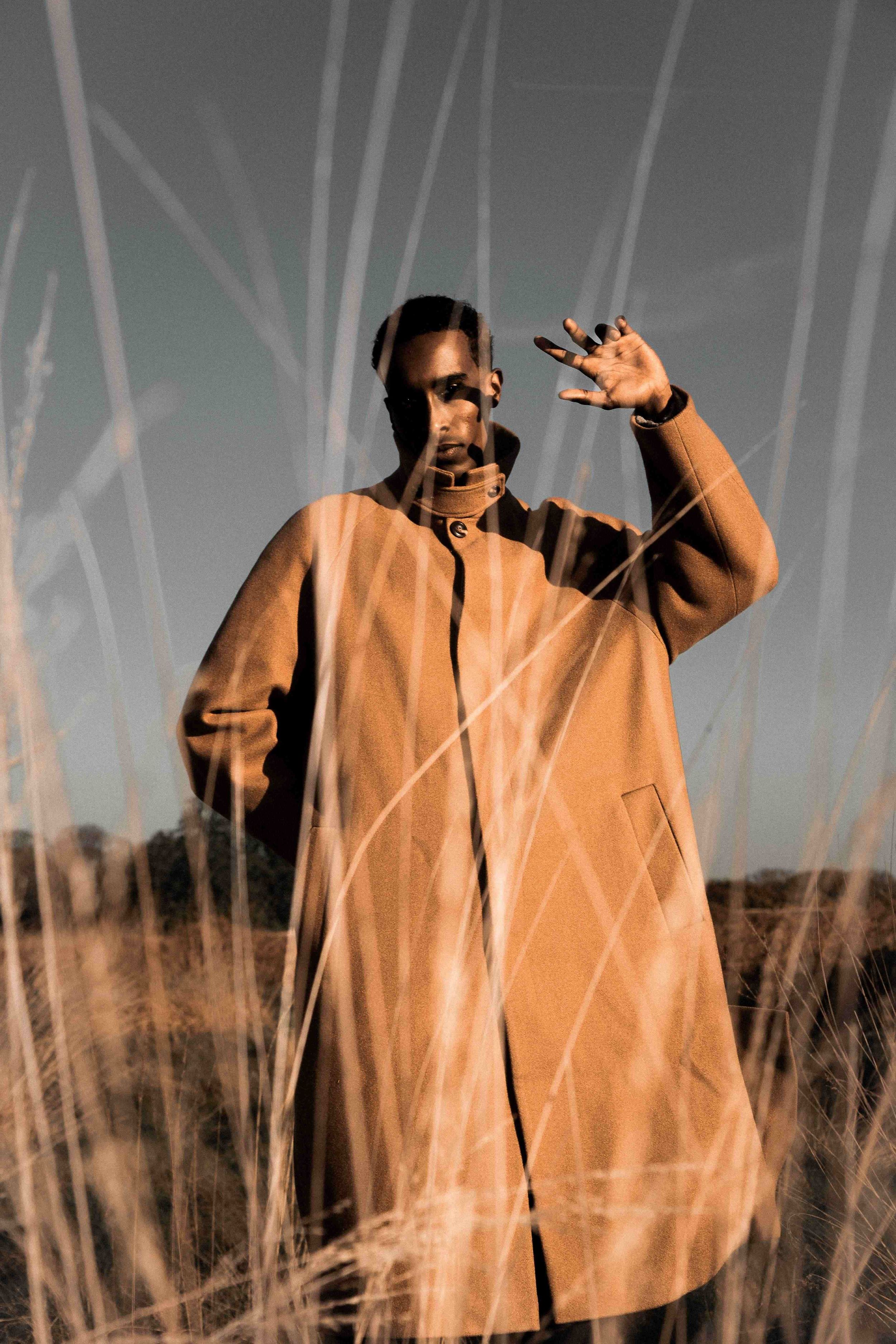 Man in tan coat standing in tall grass field, raising hand dramatically against blue sky at golden hour.