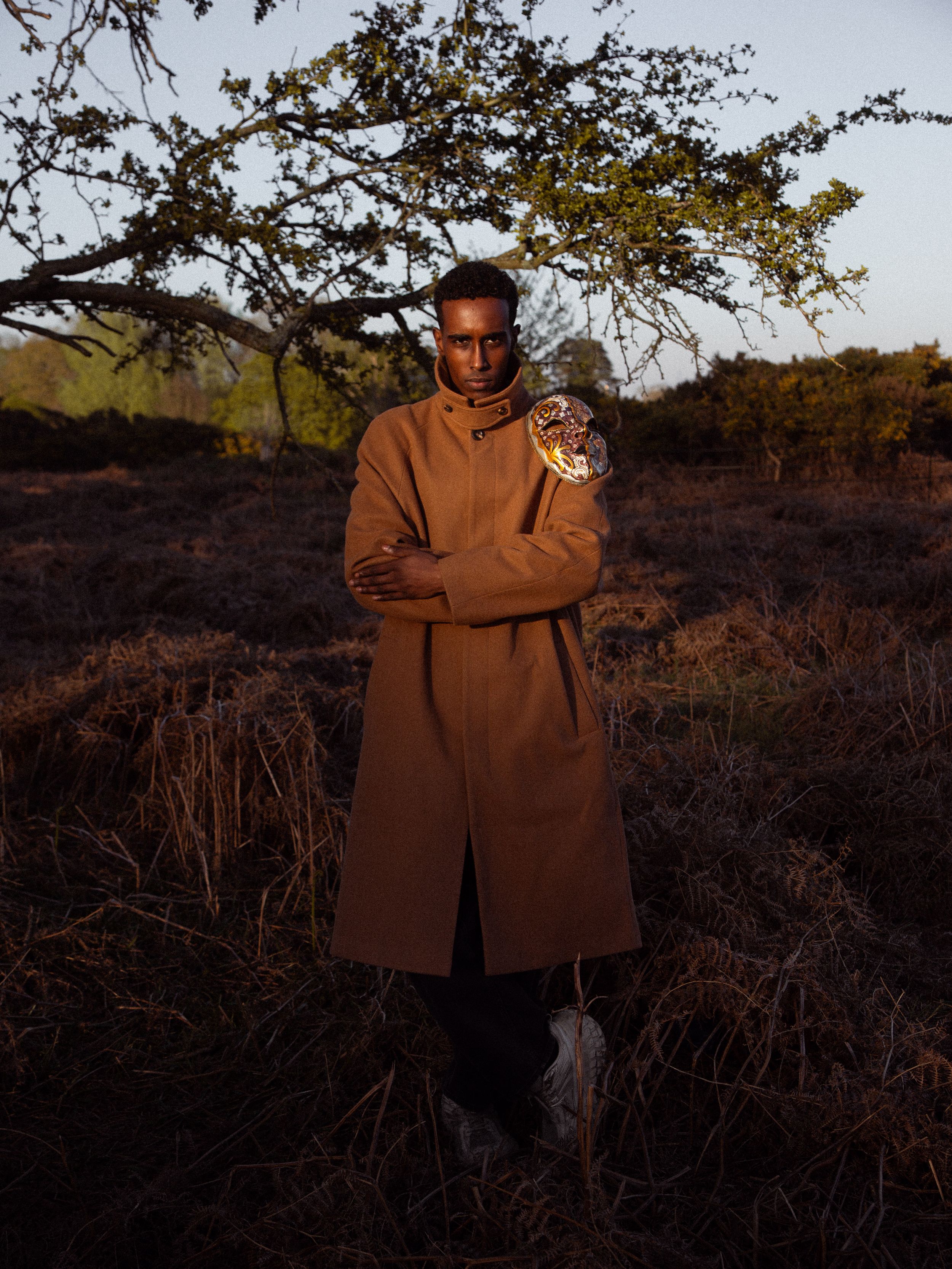 Model wearing a camel wool overcoat standing in an autumn field at golden hour, holding a decorative object.