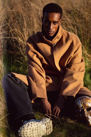 Young Black man in camel coat and dark jeans sits in golden grass field wearing white chunky sneakers.