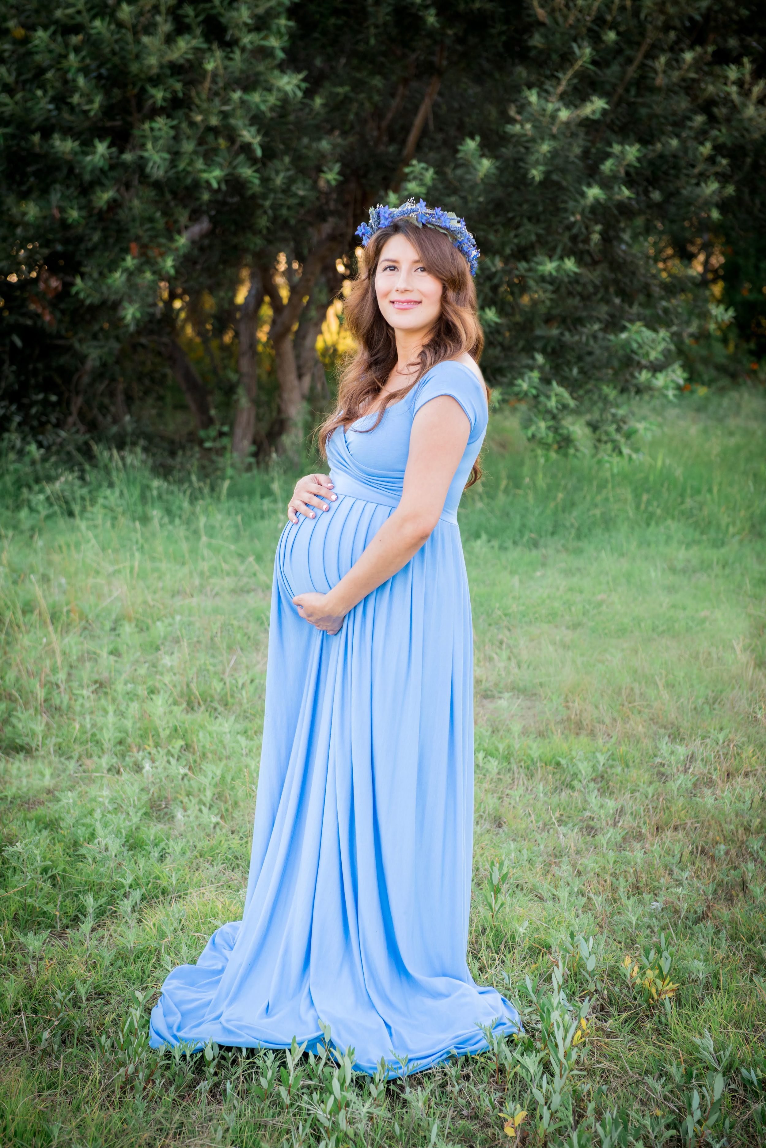Expectant mother in flowing blue dress and floral crown poses in garden setting.