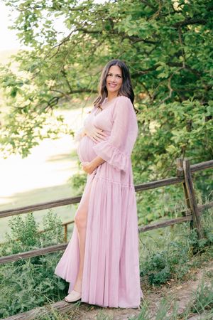 Pregnant woman in pink maxi dress posing outdoors near wooden fence with lush green trees.