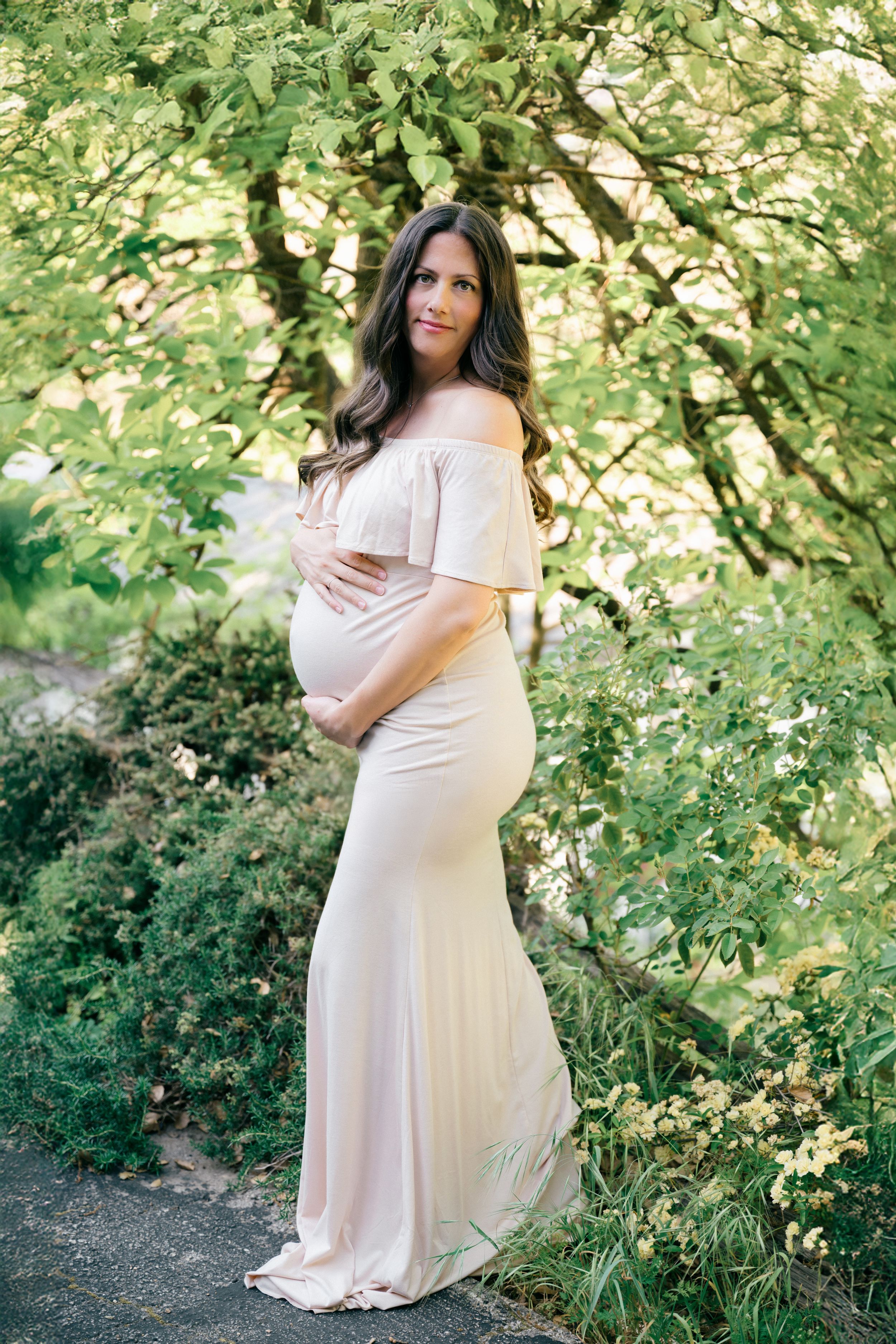 Pregnant woman in blush off-shoulder maternity gown posing outdoors among lush green trees, cradling her baby bump.