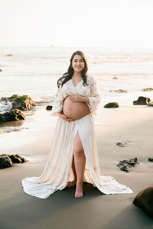 Pregnant woman in white lace maternity gown cradling baby bump on sandy beach at golden hour.