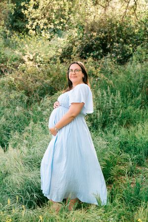 Pregnant woman in light blue dress standing in green field, cradling baby bump, wearing glasses, surrounded by lush foliage.