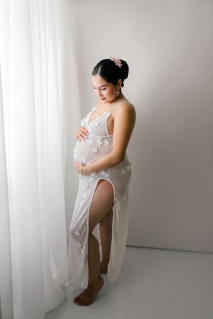 Pregnant woman in white floral lace gown standing by sheer curtains, cradling baby bump in elegant maternity photo.