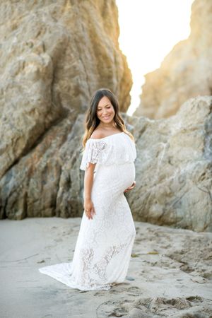 Maternity photo of woman in white lace dress standing on sandy beach near rocky cliffs.