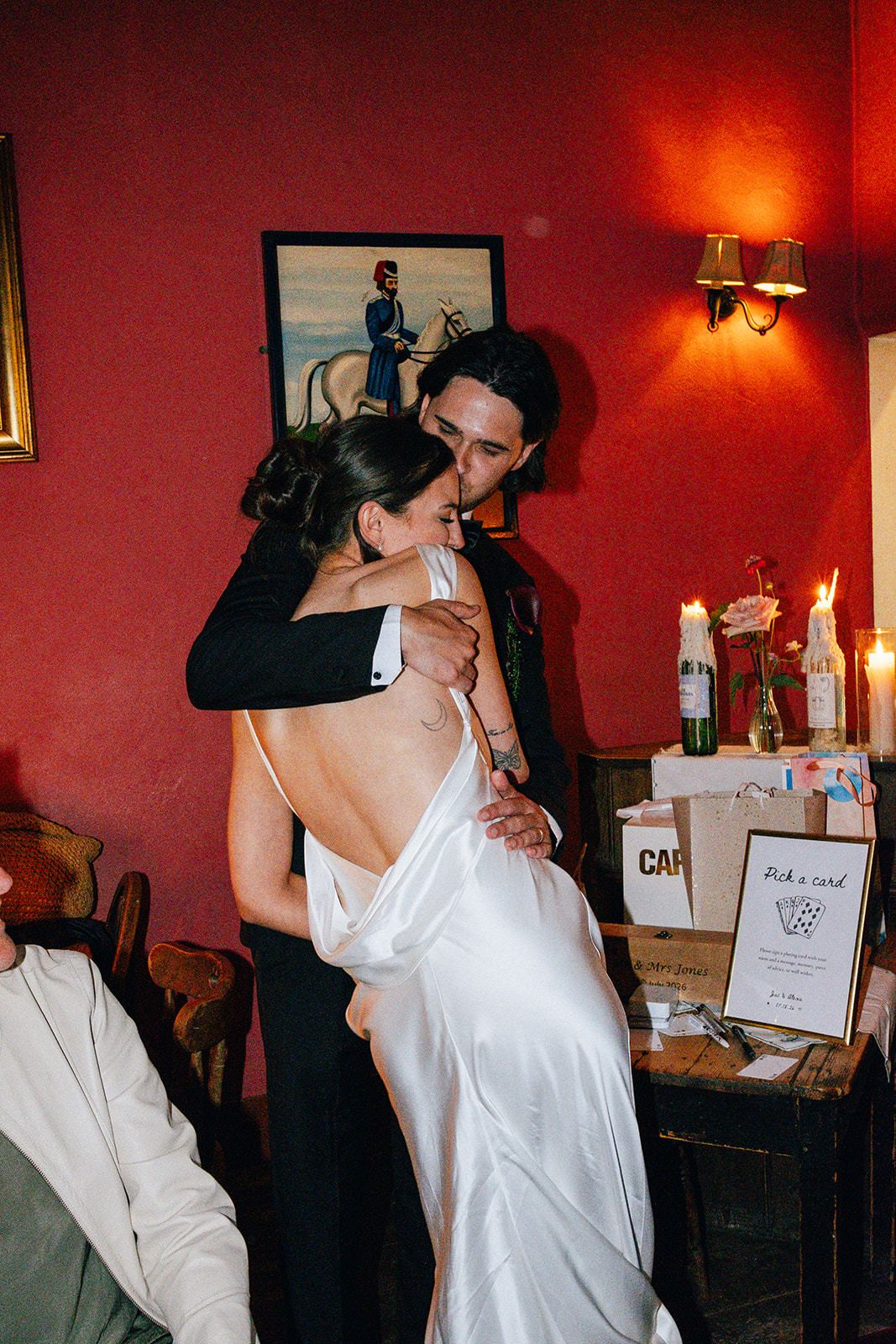 Two women share a warm hug in a rustic venue, one wearing a white wedding dress and the other a red robe.