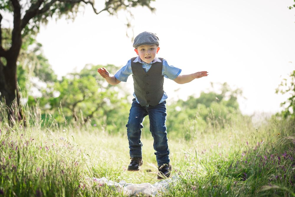 A young child in a blue newsboy cap and vest joyfully jumps in a sunny meadow on a summer day.