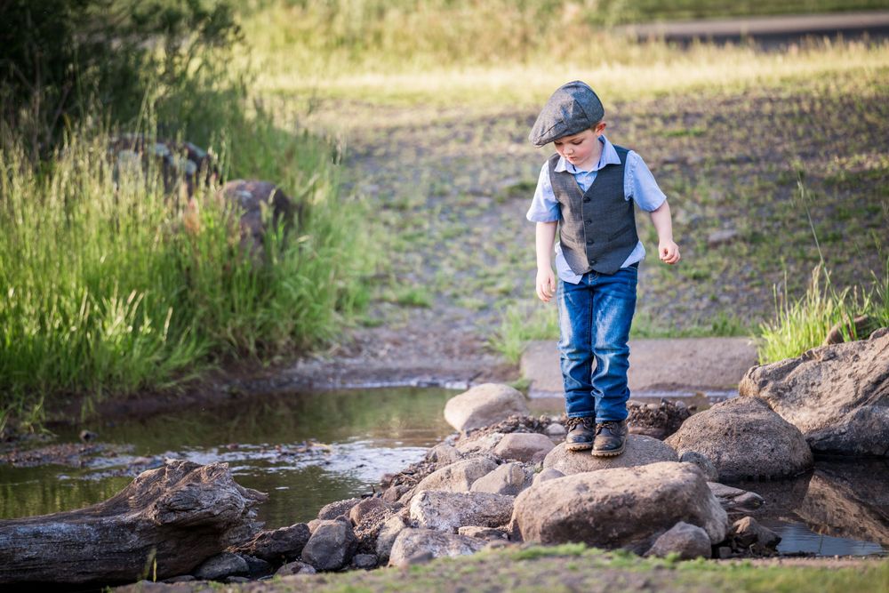 A child in overalls and baseball cap explores a rocky stream bank on a sunny summer day while wearing a backpack.