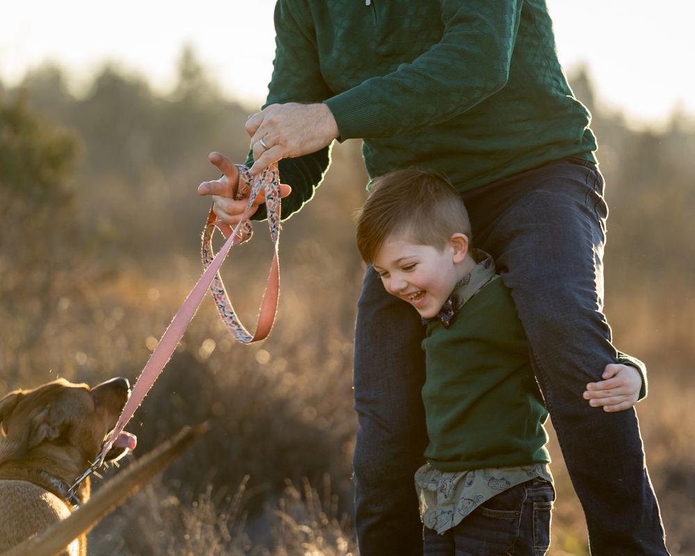 A child and parent sharing a moment outdoors with their leashed dog during golden hour.