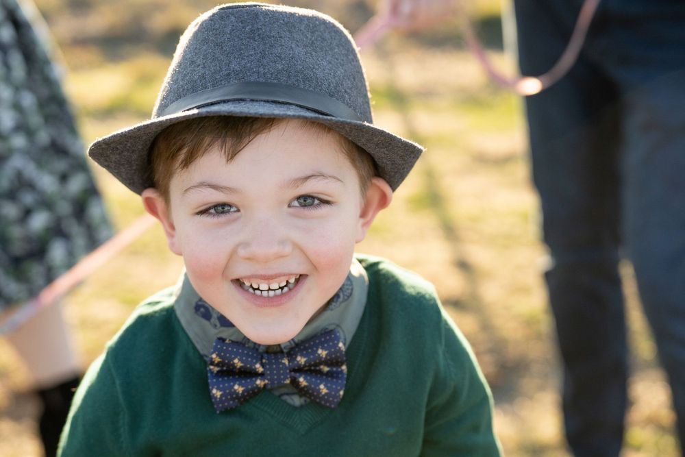 A young child wearing a gray fedora hat, bowtie and green sweater smiles warmly outdoors.