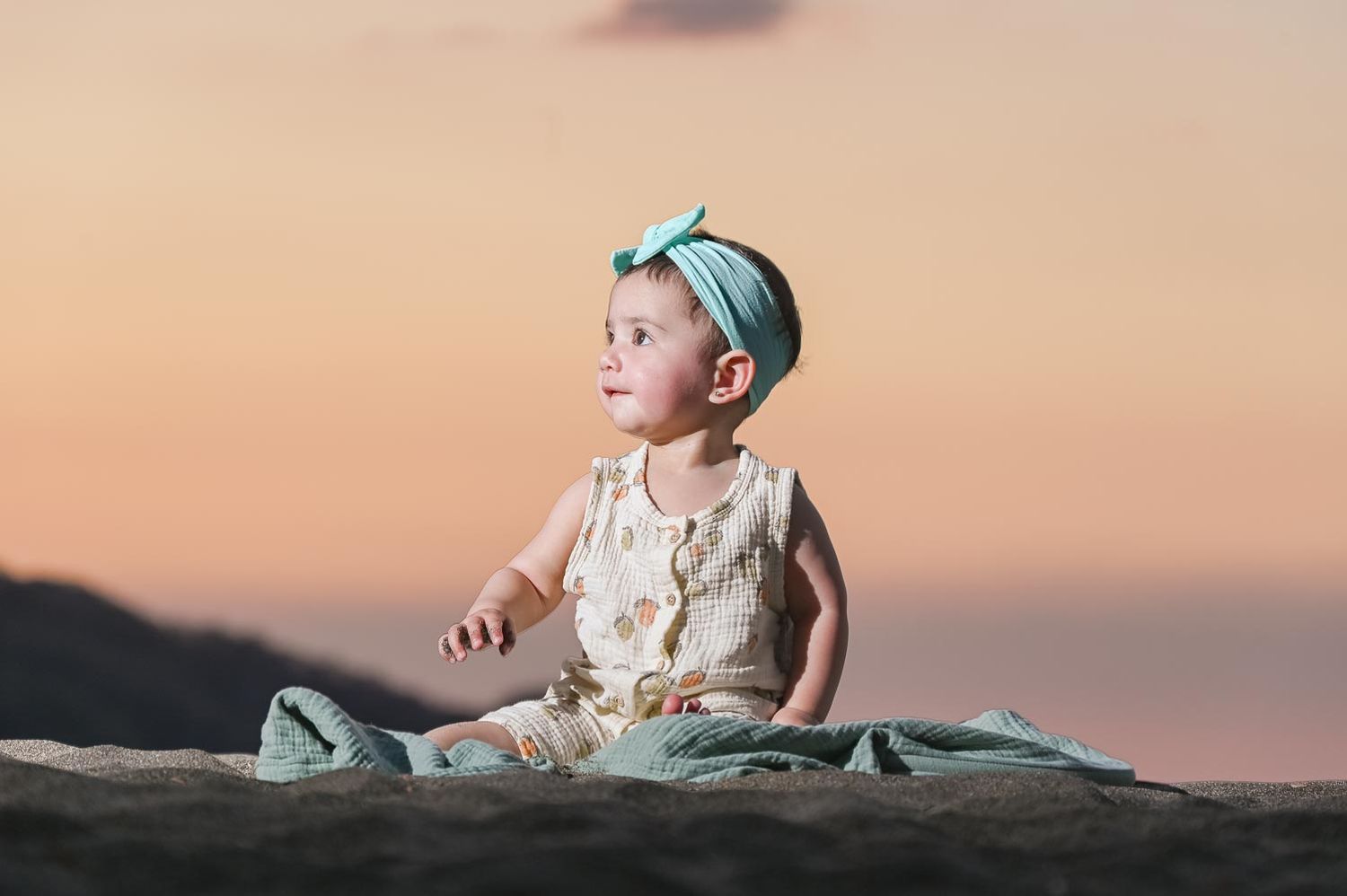 A cute baby girl in a floral romper and mint headband sits on a blanket at sunset beach.