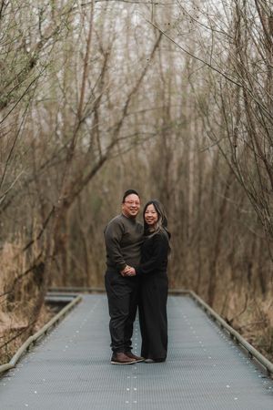 Couple smiling together on a wooden boardwalk path surrounded by bare trees in a serene outdoor setting.