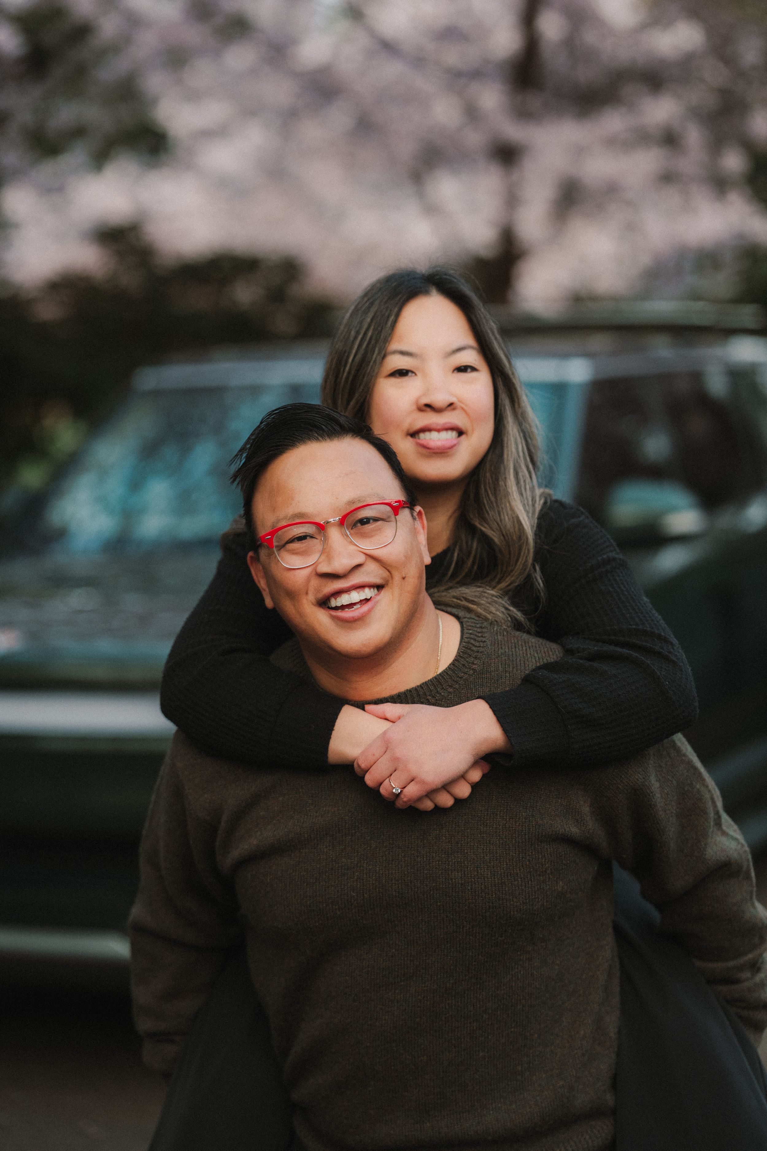 A smiling couple poses outdoors near cherry blossoms, man with red glasses giving woman a piggyback ride.