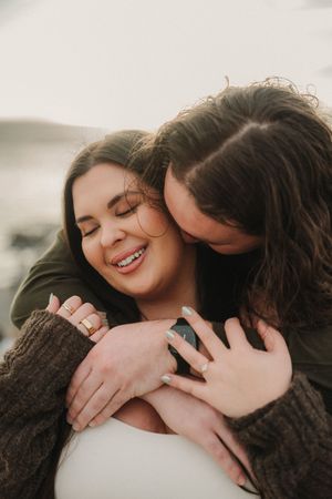 Two women sharing a tender embrace, one smiling with eyes closed as the other hugs her from behind outdoors.