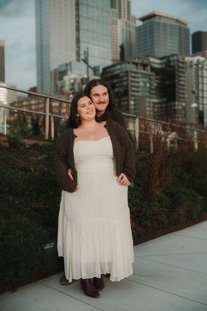 Couple posing on urban rooftop terrace, woman in white dress, man in brown cardigan, city skyscrapers in background.