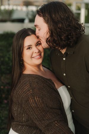 A couple shares a tender moment outdoors, man kissing womans cheek as she smiles warmly, wearing brown sweater.