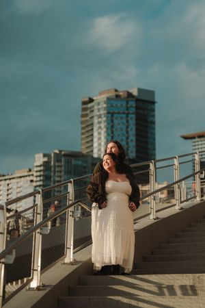 Couple posing on urban stairs, woman in white dress, man behind her, city skyline and blue sky in background.