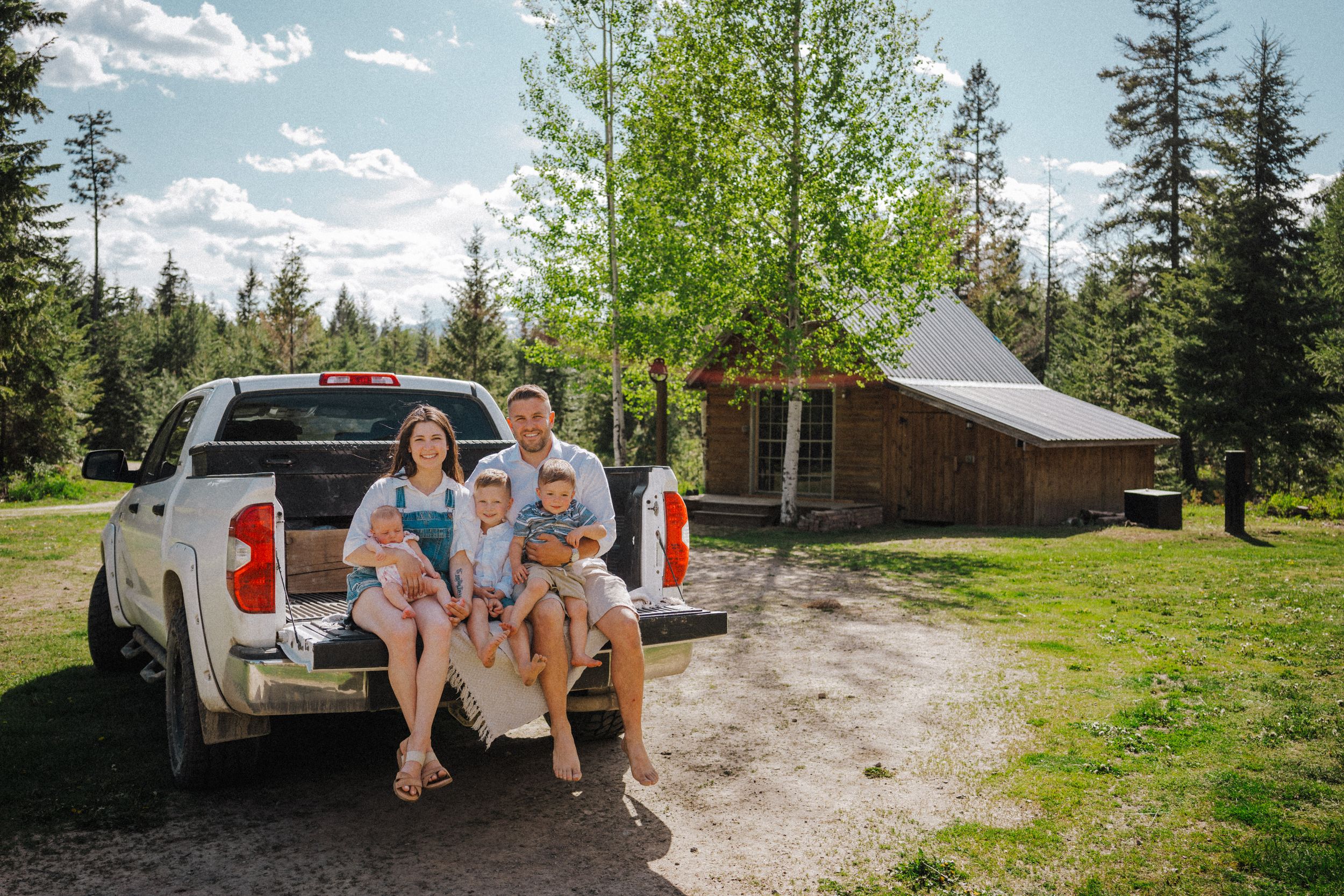 Family of four sitting on a white pickup truck tailgate near a rustic log cabin surrounded by pine trees on a sunny day.