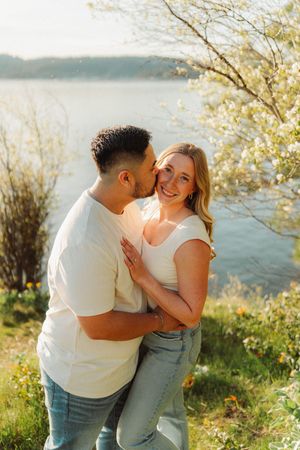 Couple embracing by a sunlit lake, man kissing woman's cheek as she smiles at camera.