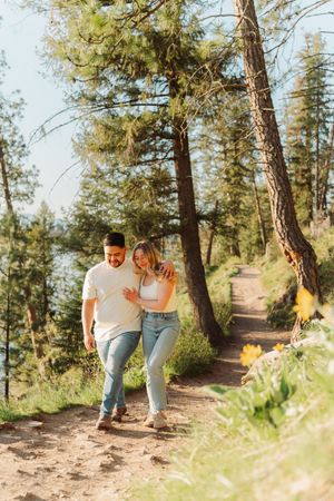 Couple walking arm in arm on a sunny forest trail surrounded by tall pine trees and wildflowers near a lake.