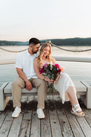 Couple sitting on a waterfront dock bench, man gifting woman a colorful bouquet of roses at sunset.