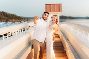 A smiling couple poses for a selfie on a deck with a snowy lake backdrop at sunset.