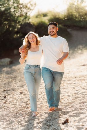 Happy couple walking barefoot on sandy beach in white tees and jeans during golden hour sunset.