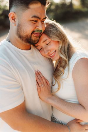 A couple shares a tender embrace outdoors, with the woman smiling and resting her head on his chest.