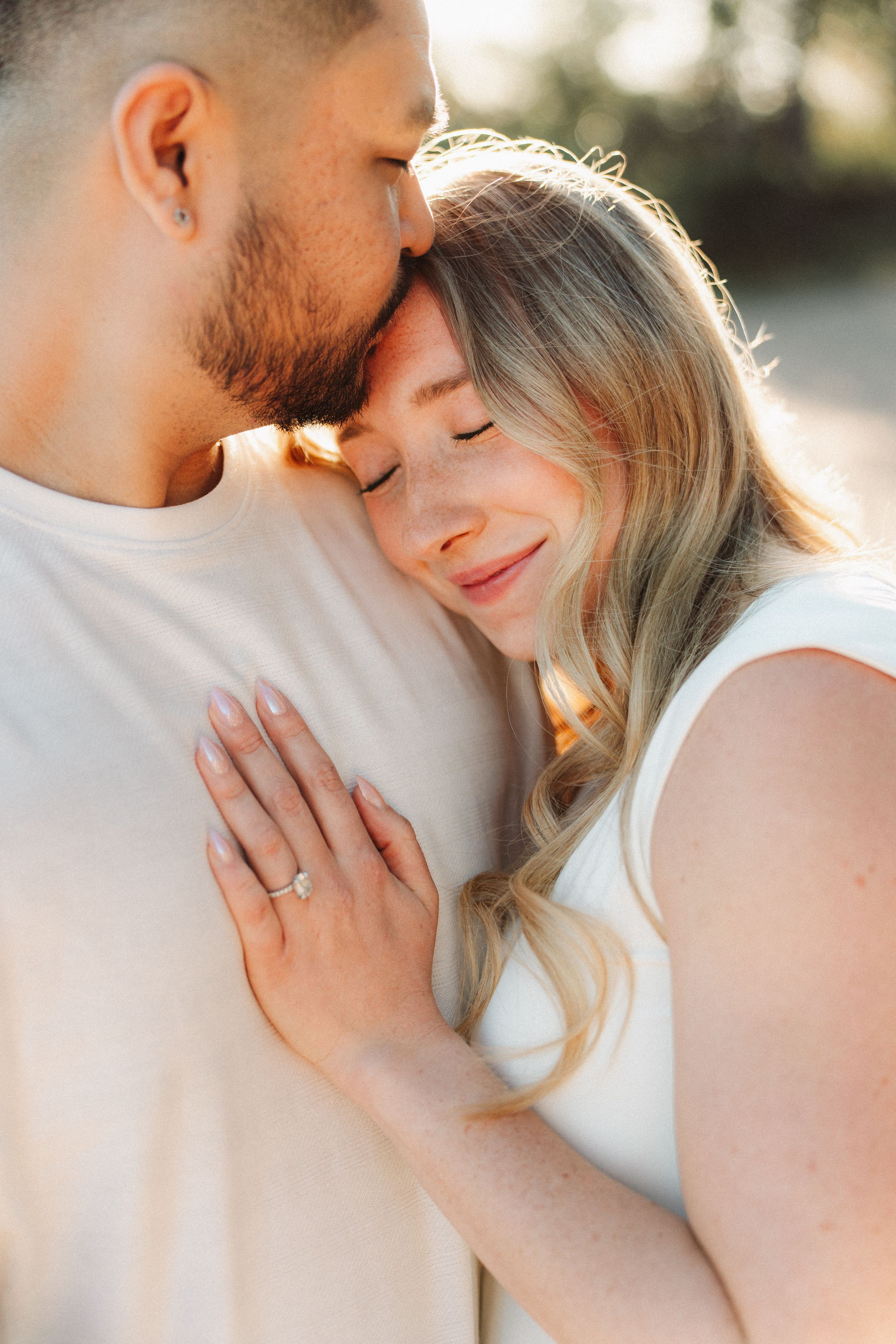 Couple sharing tender moment, woman with engagement ring resting head on mans chest outdoors.